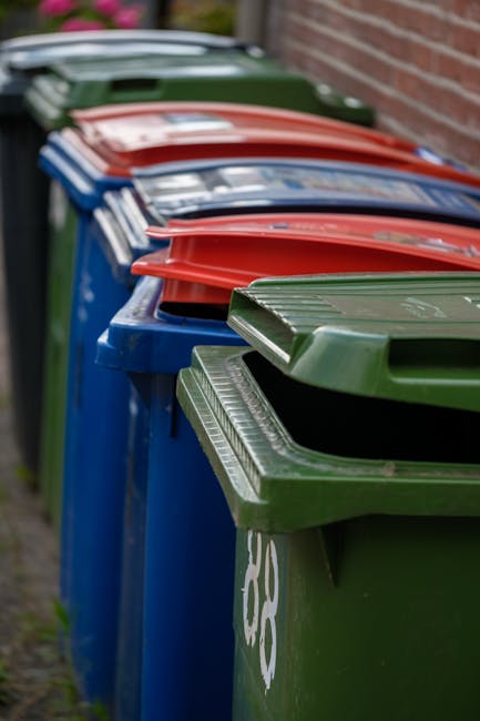 A row of five large wheelie bins placed on a paved driveway next to a brick wall, with the closest bin being green and the others in blue, red, and dark green. The bins have textured, ribbed lids with